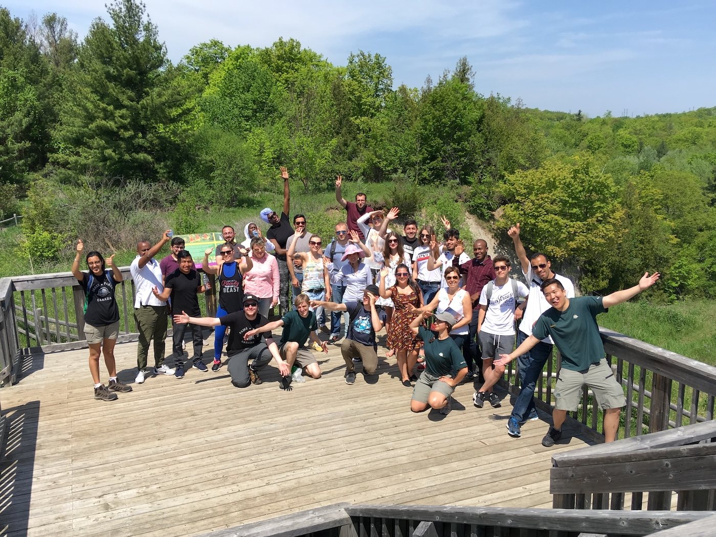 A large group waving from a wooden boardwalk lookout above a green valley, with Parks Canada staff among them