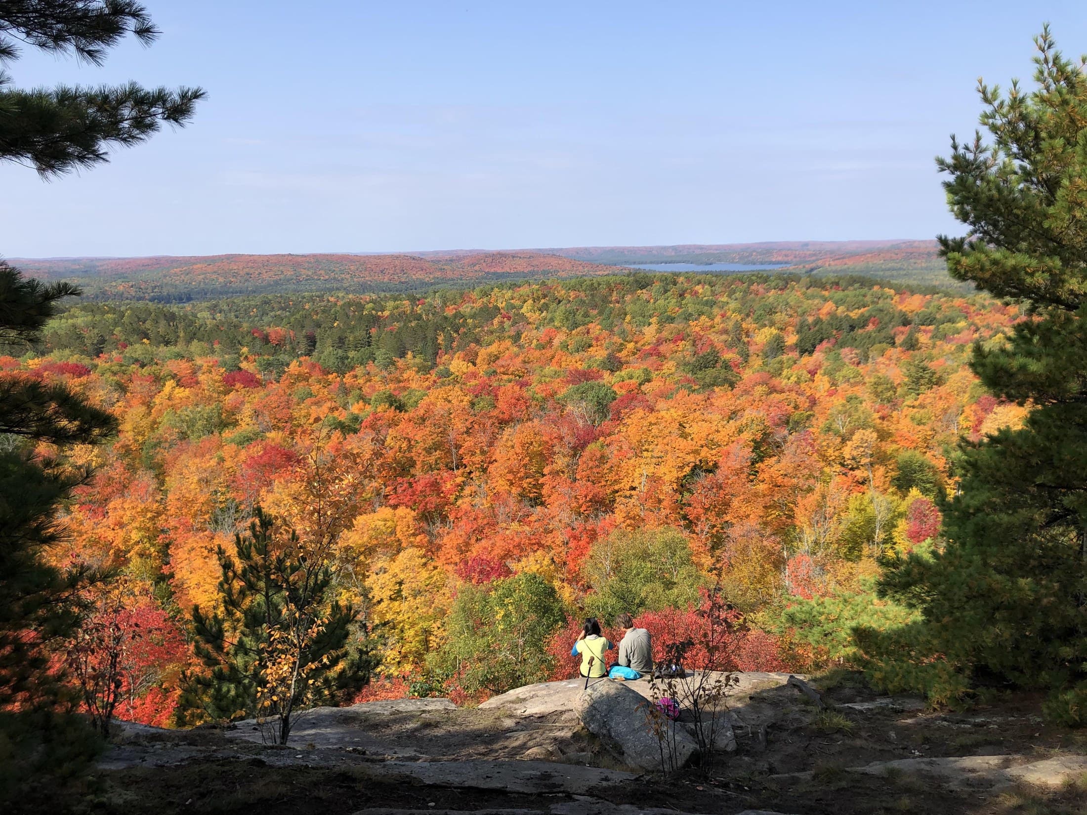 Two Parkbus riders sitting on a rock at a lookout above a valley of red, orange and gold maple in Algonquin, 26 September 2026