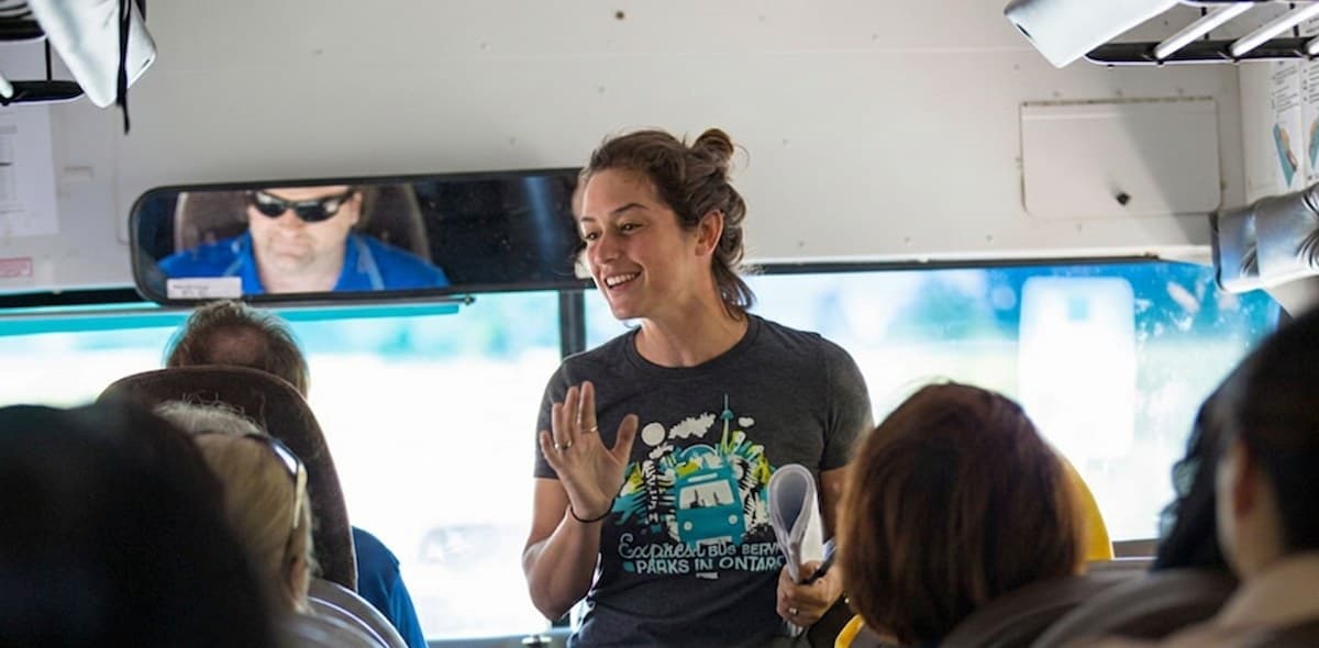 A Parkbus trip ambassador standing at the front of the bus in a Parkbus shirt, talking to the passengers